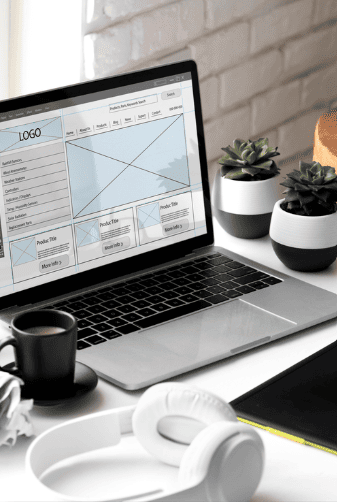 Imagem de um computador em cima de uma secretária, com headphones, plantas e uma caneca. No pc vê-se o layout da construção de um wireframe